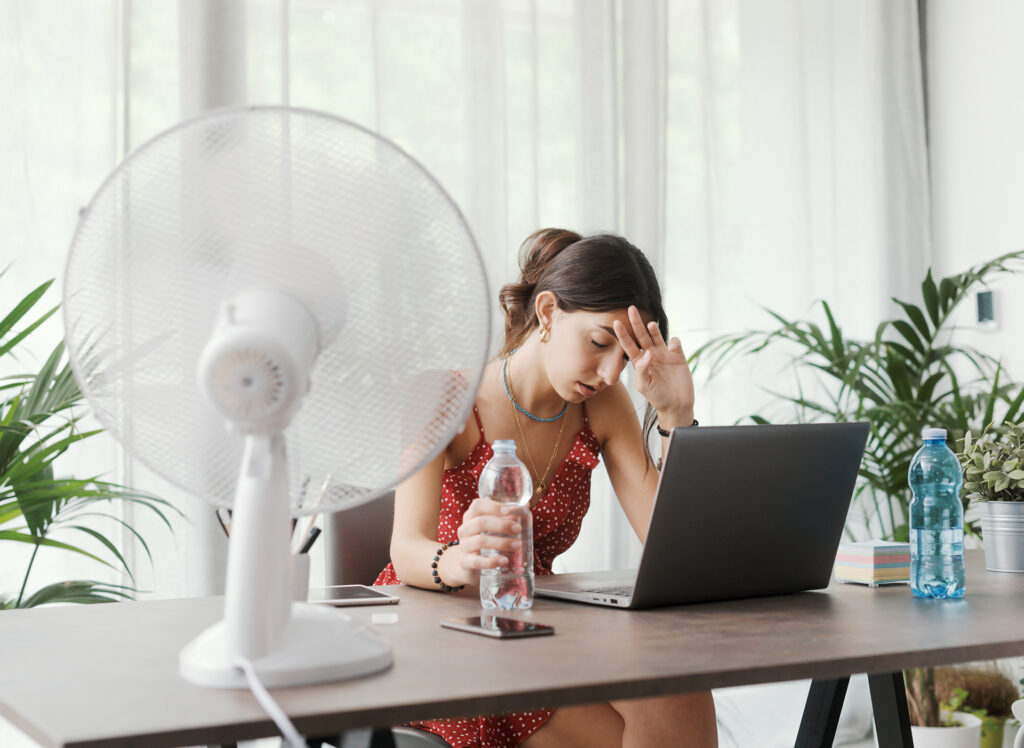 A woman at her desk in Rainbow Lakes Estates, fanning herself with a paper because her air conditioner isn't working, a sign that she needs air conditioner repair.